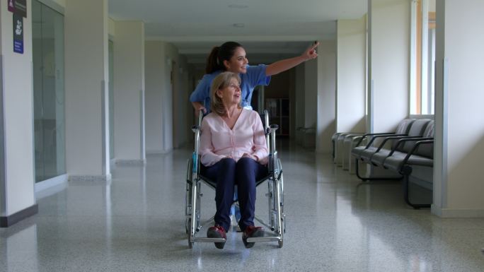 友好的护士走过医院走廊，带着一位坐在轮椅上的老年女性患者，一边指着一边给她看东西