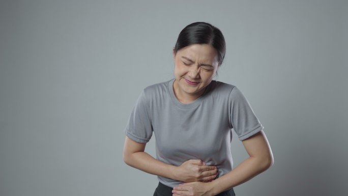 一位亚洲女性因胃痛而病倒，她站在灰色的背景上，独自一人。