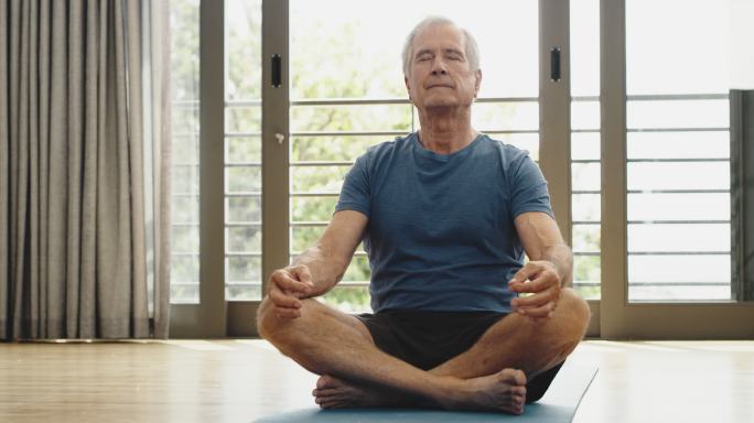 一位老人坐在家里练瑜伽。老人坐在垫子上冥想