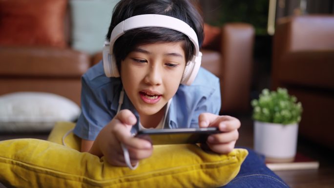 令人心寒的周末假期，电子竞技游戏在线，亚洲男孩戴着耳机，手在网上玩智能手机游戏，兴奋而有趣地躺在家里