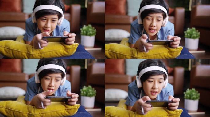 令人心寒的周末假期，电子竞技游戏在线，亚洲男孩戴着耳机，手在网上玩智能手机游戏，兴奋而有趣地躺在家里