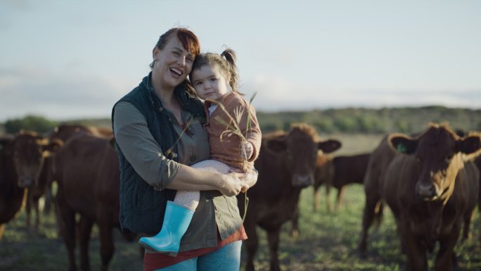 母女俩在农场里的画像，靠近他们的奶牛。农村单身母亲微笑着和她的小孩站在一起，手里拿着干草，对着相机挥