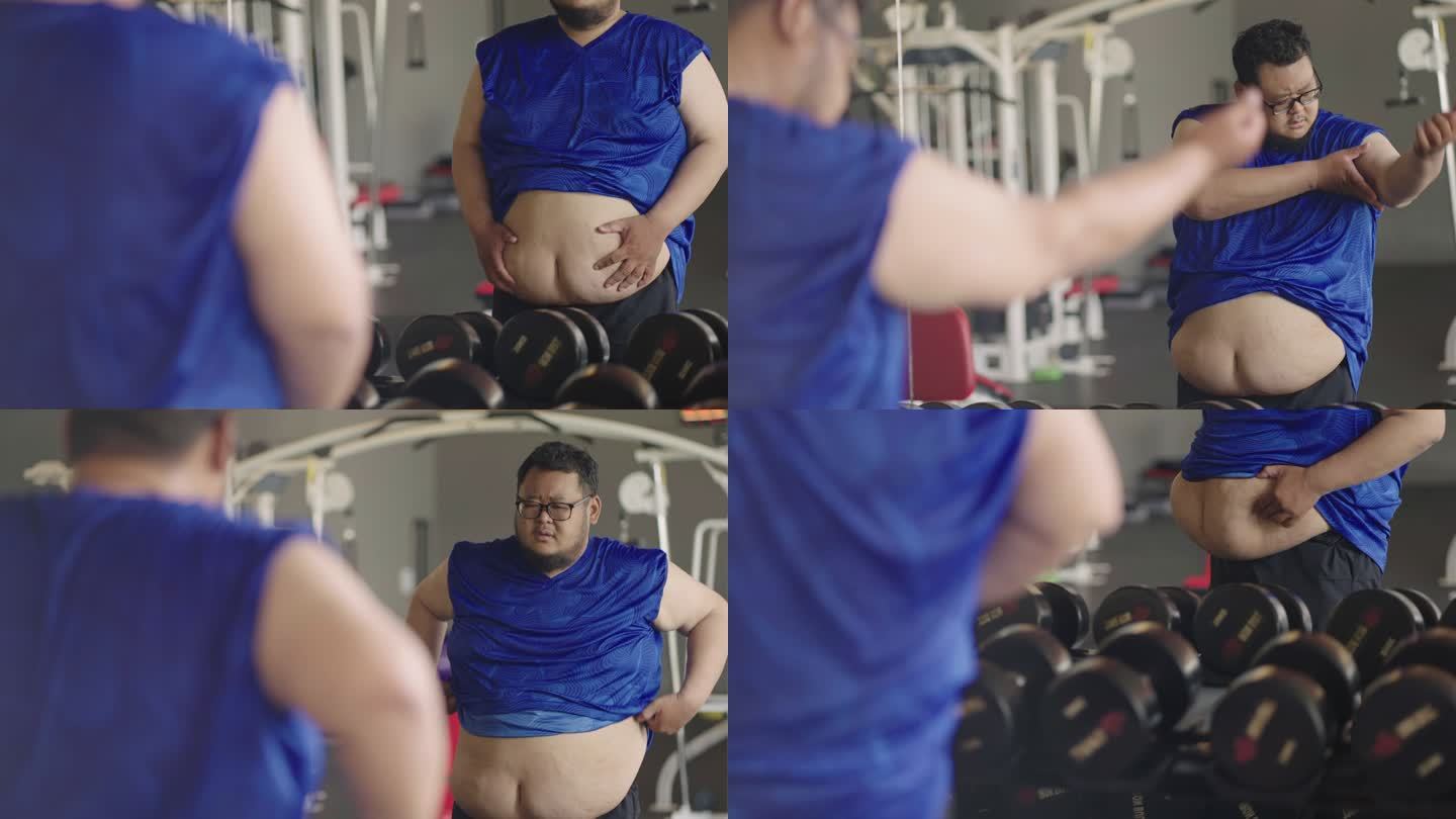 一个非常胖的男人站在镜子前看着自己，拉起衬衫，在室内健身房里摇晃着他的大肚皮和前臂。