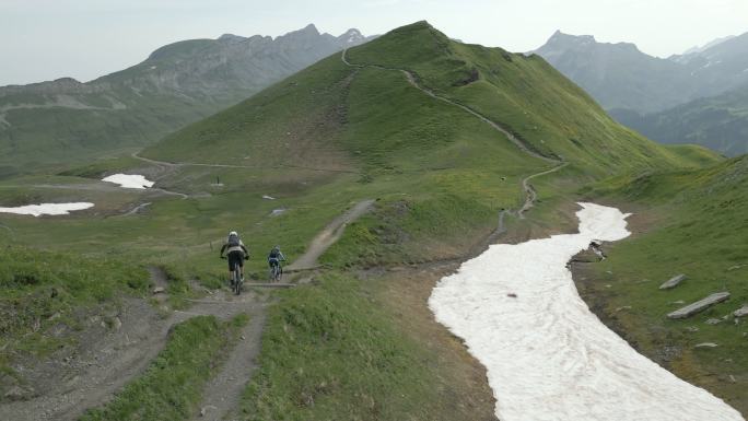 日出时山地自行车手穿越高山草甸小径的鸟瞰图