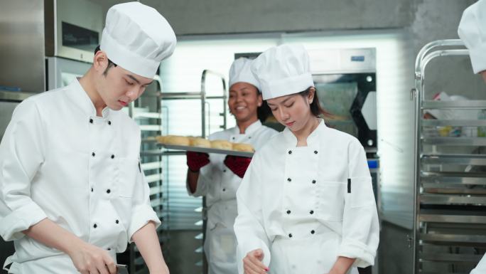 穿着白色厨师制服的快乐的多种族年轻成人和高级亚洲男面包师在商业厨房一起制作面包、面包和法式面包。非洲