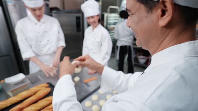 高角度视角：身着白色厨师制服的多种族年轻成人和高级亚洲男面包师在商业厨房一起制作面包、面包和法式面包