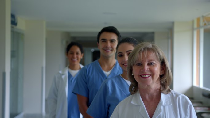 不同医学专业的快乐团队站成一排，面对镜头微笑着站在走廊的医院里
