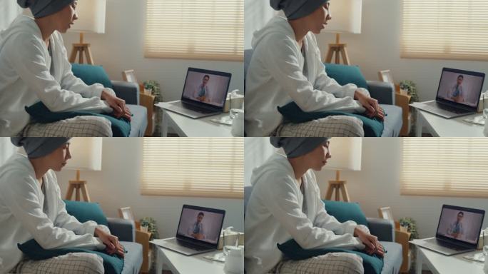 一位患有癌症的亚洲年轻女性使用笔记本电脑与坐在家里客厅沙发上的医生交谈。