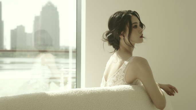 这位身着白色连衣裙的美丽亚洲新娘坐在落地窗前的沙发上慢镜头拍摄