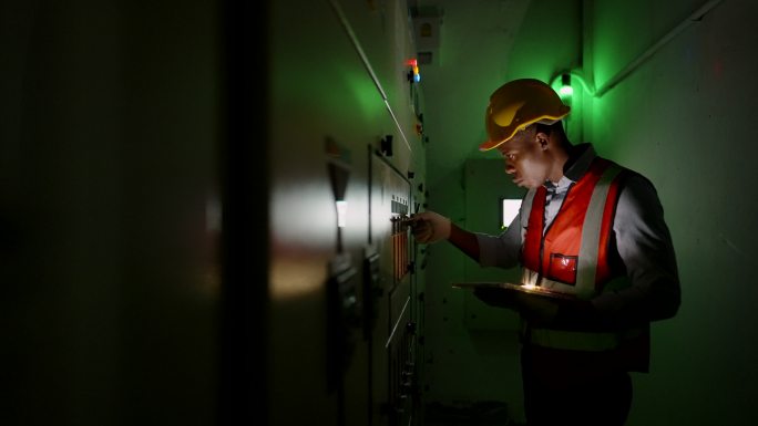 非裔美国工业工程师在夜班时穿戴个人防护装备，在工厂控制室使用平板电脑检查制造公用系统的能源生产过程的