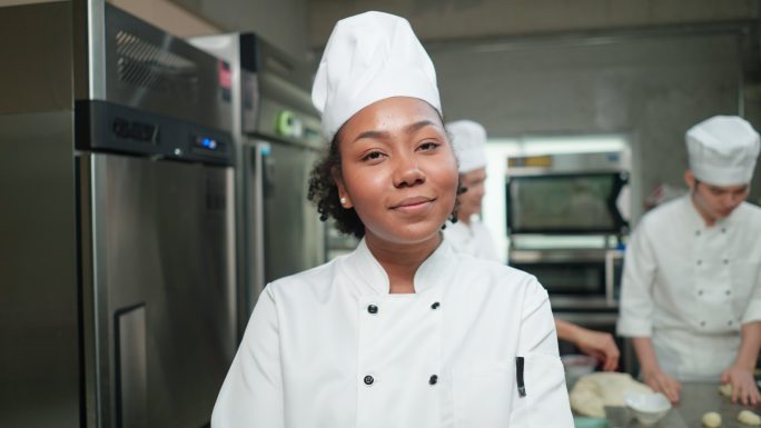 肖像：快乐的非洲年轻成年女厨师，身着白色厨师制服，微笑着，站着，交叉双臂，手持木制擀面杖，其他厨师为