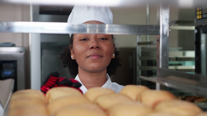 头像：快乐的非洲年轻女性面包师，身穿白色厨师制服，戴着烤箱手套，在面包店手推车架上检查刚出炉的面包，