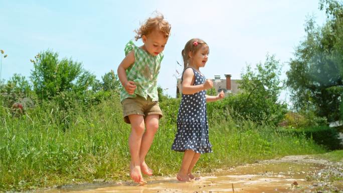 两个赤脚的小女孩在阳光下的泥潭里跳上跳下