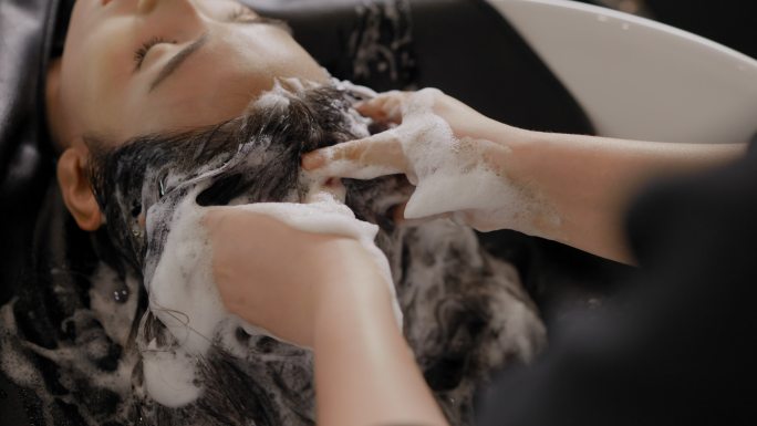 在一家美容院，一位放松的亚洲女性顾客斜靠在浴缸上，闭上眼睛，理发师用淡淡的香草洗发水轻柔地按摩她的头