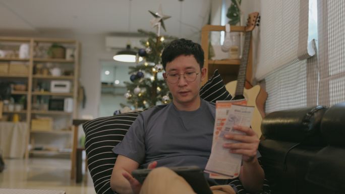 一位亚洲家庭男子在沙发上放松，通过在线支付、电费和水费来支付家里的账单。