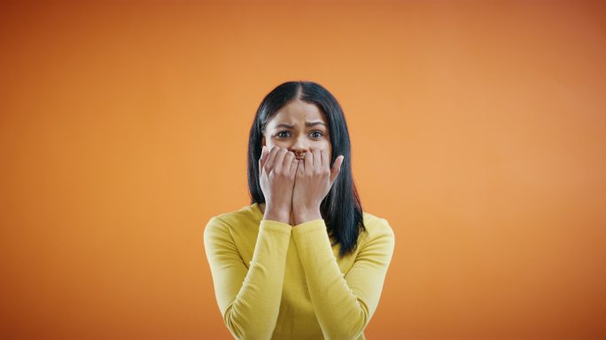 惊恐的年轻女子在橙色背景下把手放在脸上。混血女性用双手捂住嘴，露出恐惧的表情