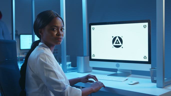 一位年轻的黑皮肤实验室医生，穿着白色实验服，在她的工作场所，在一个现代化实验室的电脑显示器上，转向一