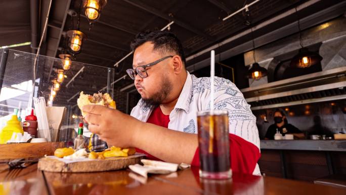 胖子年轻人在服务员端汉堡包之前使用手机的情景。男人在快餐店饥肠辘辘地吃汉堡包。