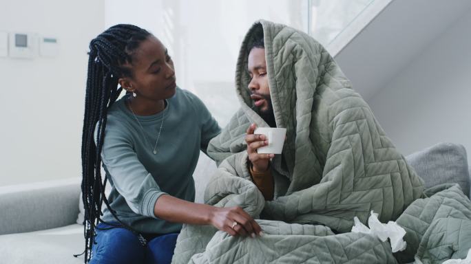 一位年轻女子在家照顾患流感的丈夫。女人帮助生病的男人，用毯子盖上，用纸巾擤鼻涕