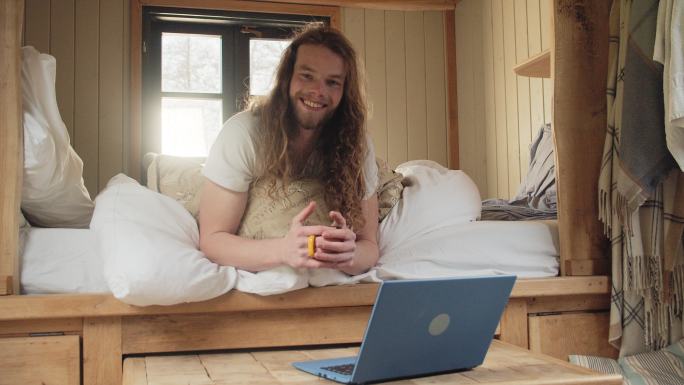 在小屋的小床上用笔记本电脑画一个快乐的年轻人，他喝着咖啡