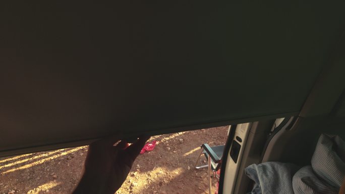 一名男子坐在他的货车里，打开门前的灰色卷帘，伸开双腿，他的妻子躺在户外的吊床上，看着她的手机，背景是