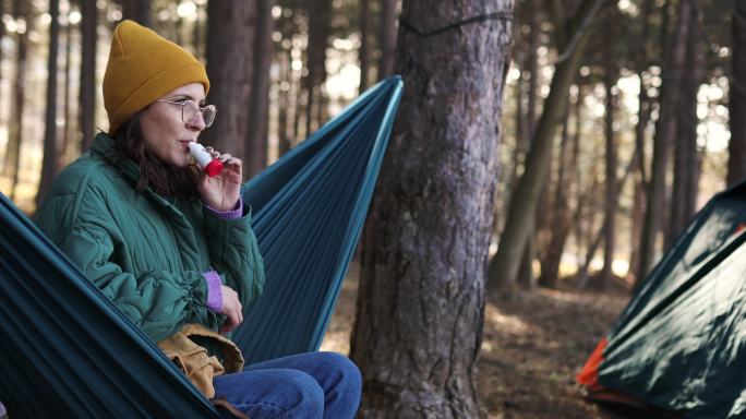 一位千禧年女性躺在树林中的吊床上，使用哮喘吸入器