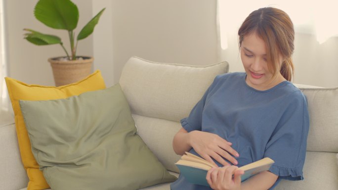 快乐的亚洲孕妇在家看书，轻轻抚摸肚子，怀孕/分娩