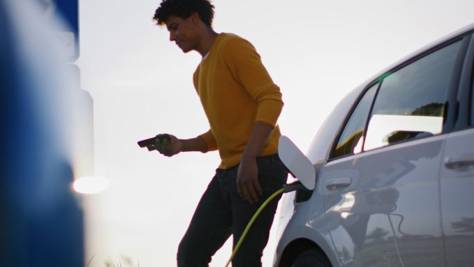SLO MO Mulatto年轻男子在为电动汽车充电时使用智能手机