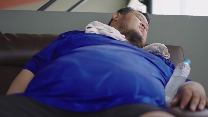 在健身房锻炼后，一个体重超标的家伙躺在沙发上，胳膊上拿着水瓶。