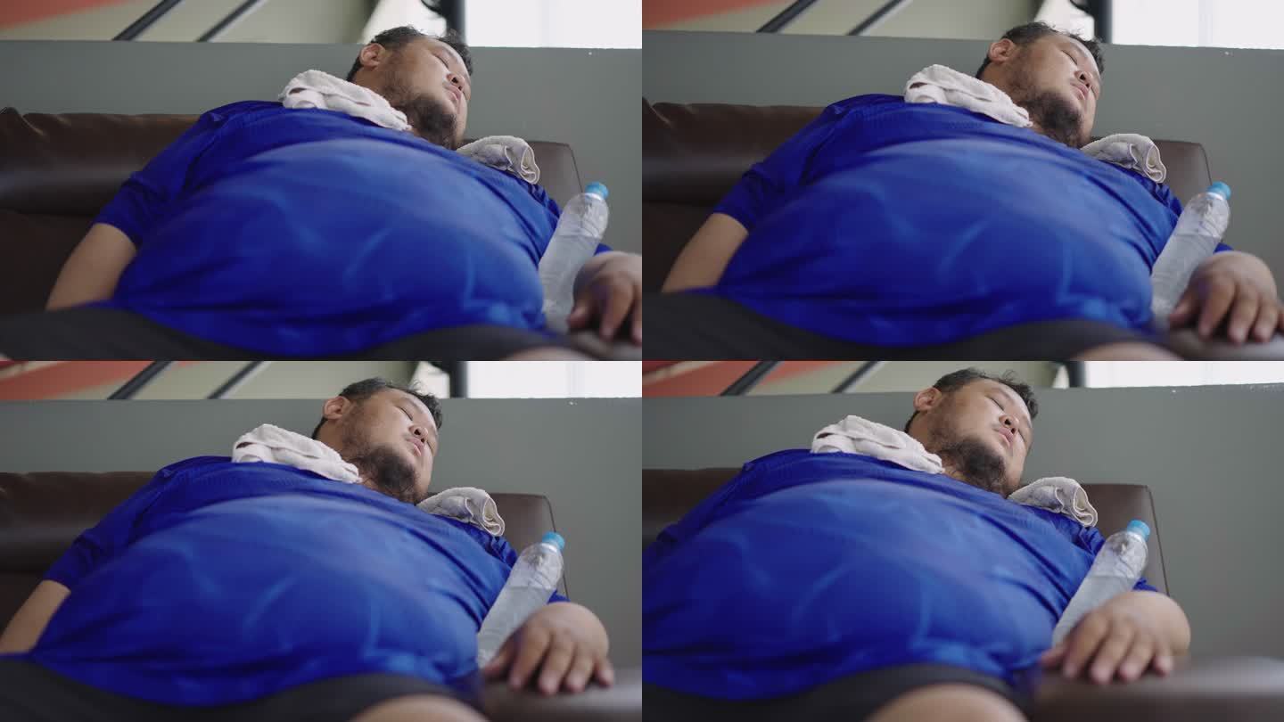 在健身房锻炼后，一个体重超标的家伙躺在沙发上，胳膊上拿着水瓶。