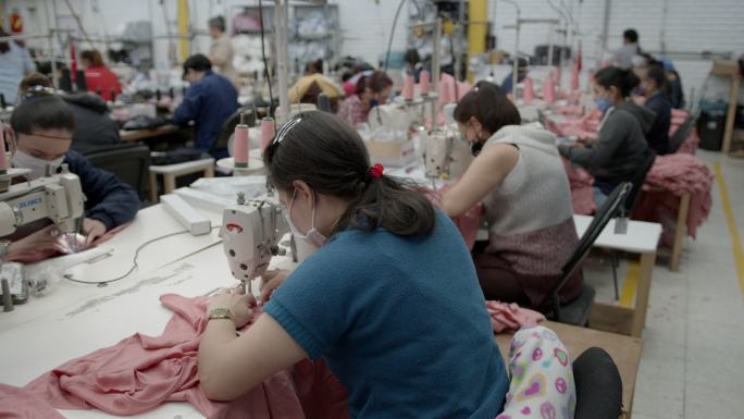 拉丁美洲妇女在服装厂的缝纫站工作，每个人都戴着防护口罩