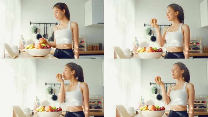 穿着运动服的亚洲活跃美女在厨房吃苹果。年轻漂亮的女孩心情愉快，喜欢吃水果和健康食品，在家里节食减肥。