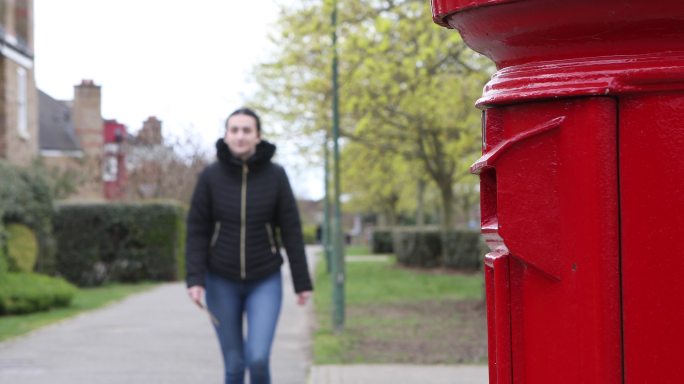 一位年轻女子走在人行道上，走向一个红色的邮箱，并贴了一封信