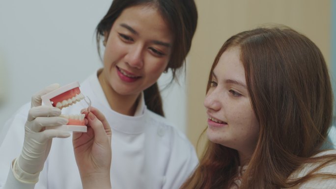 年轻的女牙医和十几岁的女孩在牙科诊所的牙医练习室拿着模型牙和牙线。