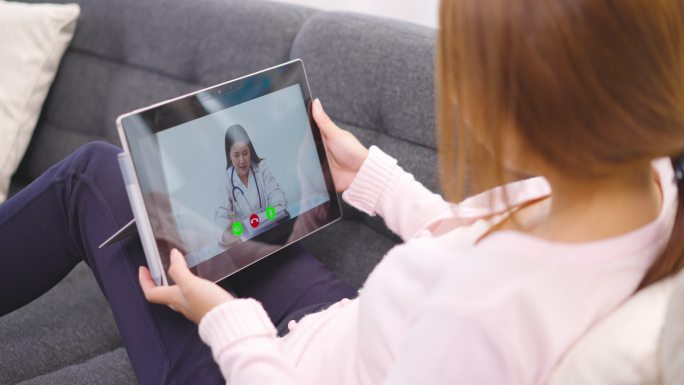 亚洲年轻女性躺在沙发上，与医生在家里的平板电脑上进行视频会议