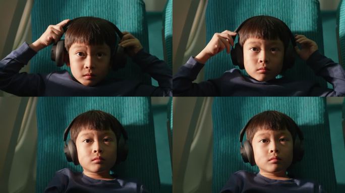 一个男孩坐在一架商用飞机旁边的窗户上，他举起耳机戴上它。为了在旅途中放松，享受乘坐飞机旅行的兴奋。