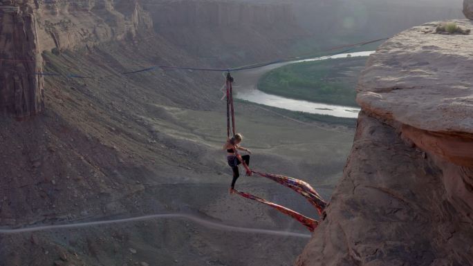 杂技青年女子在犹他州摩押附近的一个峡谷上空的松弛线表演空中丝绸，背景是黄昏时的绿河峡谷