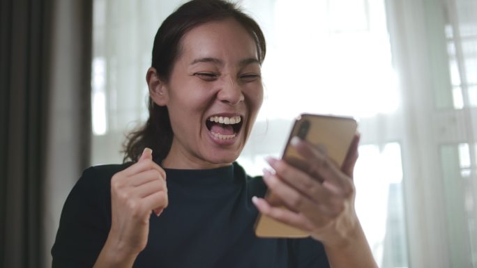 年轻的亚洲女性手持智能手机，庆祝获奖或欢呼手机获奖