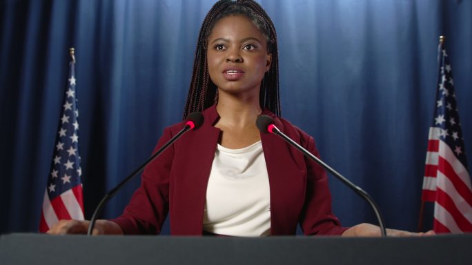 一位身穿红色夹克的年轻非裔美国政治演讲者站在蓝色幕布上，举着美国国旗发表演讲