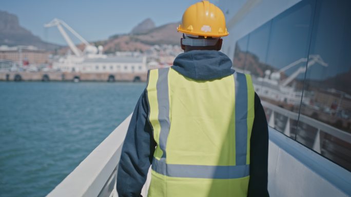 一名造船修理工戴着安全帽、反光背心和个人防护装备工作服在货船上行走的后视图。作为一名机械师在海洋上工