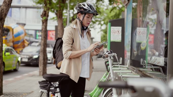 一位亚洲美女在自行车公共停车场用智能手机打开公共自行车的头盔，另一位女士正在注册租用公共自行车