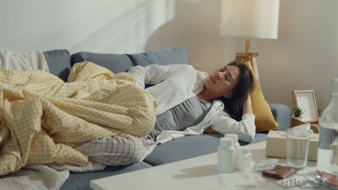 一位患有严重腹痛的亚洲年轻女性躺在家里客厅沙发上的毯子下。