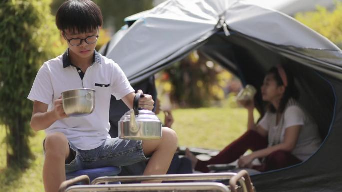亚裔中国青少年坐在后院的露营帐篷前泡茶。他的家人坐在营地里，