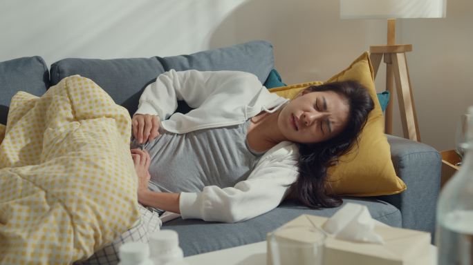 一位患有严重腹痛的亚洲年轻女性躺在家里客厅沙发上的毯子下。
