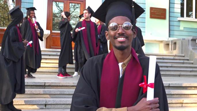 一个身着毕业礼服和迫击炮的男人，手里拿着毕业证书，对着镜头微笑