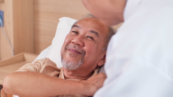 后视图，男护士医生在医院探望躺在床上的60岁亚洲老人，一边抚摸他的肩膀，一边聊天和安慰。医护人员共同