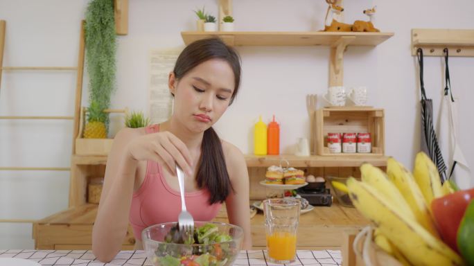 穿着健身房衣服的亚洲女性对节食不满意，吃蔬菜时脸色沉闷。