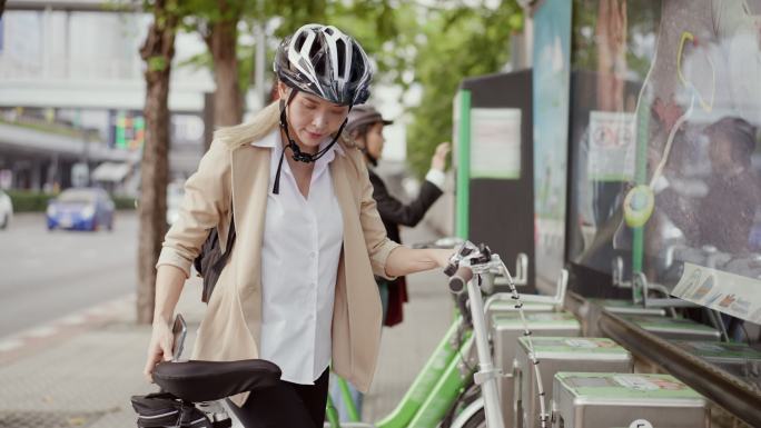 市中心有公共自行车租赁服务。一位戴着头盔的亚洲女商人正在使用手机解锁和骑自行车，另一位女商人也在使用