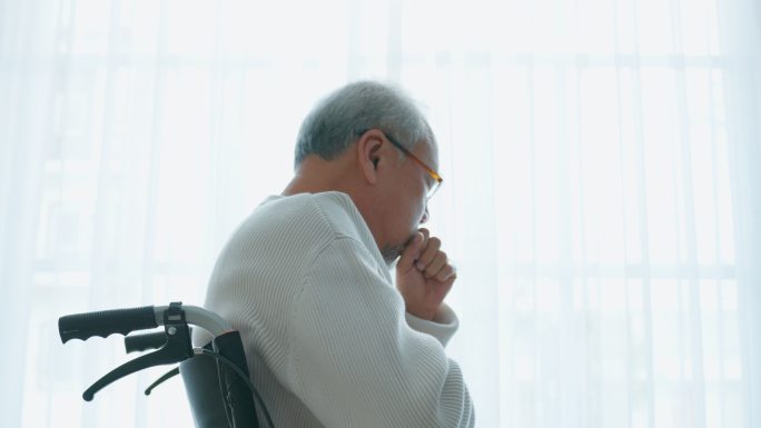 亚洲老年男性独自坐在轮椅上，退休后留在家里。不开心的老年残疾人咳嗽，担心医疗问题。医院医疗保健和医疗