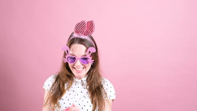 美丽的年轻女子戴着小兔子耳朵和粉色太阳镜，在手指上旋转复活节彩蛋。
